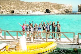 Skupina studentů mává vedle lodi v Blue Lagoon, Comino