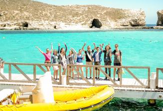 Skupina studentů mává vedle lodi v Blue Lagoon, Comino